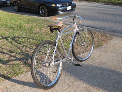 Chrome bicycle with leather seat and brown grips parked on sidewalk next to a dark blue car