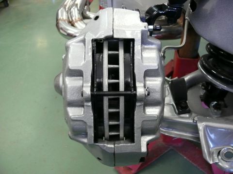 Close-up of a silver brake caliper with brake pads visible. Exhaust headers are in the background