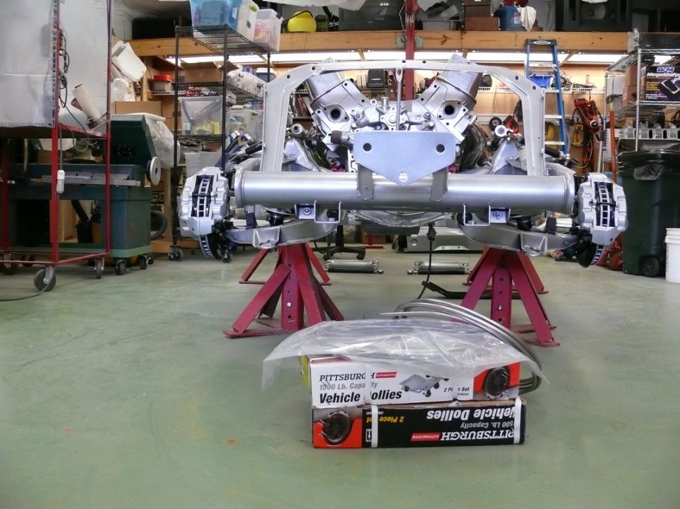Car chassis on red jack stands with V8 engine and suspension components. Boxes of vehicle dollies are in the foreground