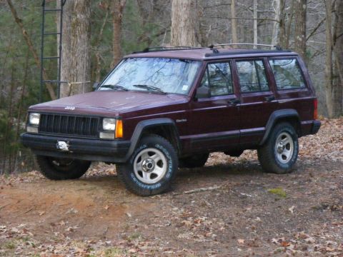 Maroon Jeep Cherokee XJ parked on a dirt path in woods. Has a ladder on a tree behind it