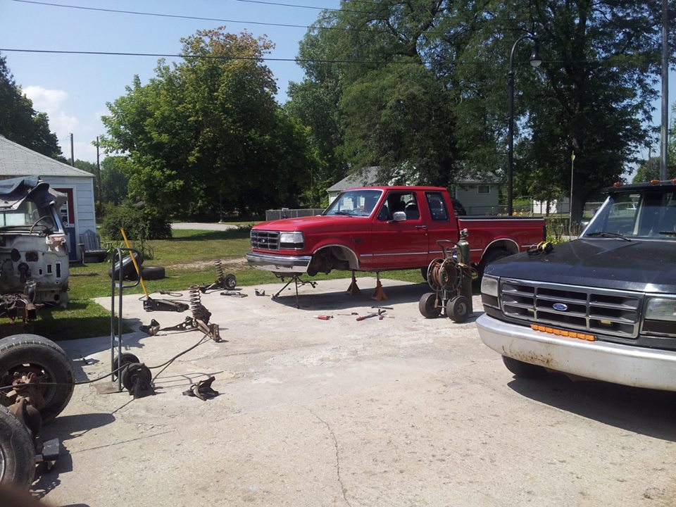 Red Ford F-Series pickup truck from the mid-1990s on jack stands, front end disassembled. Black Ford pickup in foreground. Parts and tools scattered on concrete