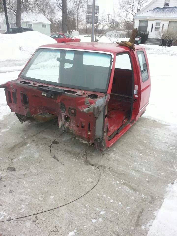 Red truck cab shell sitting on a concrete driveway in the snow. The front end is missing body panels and the engine bay is exposed