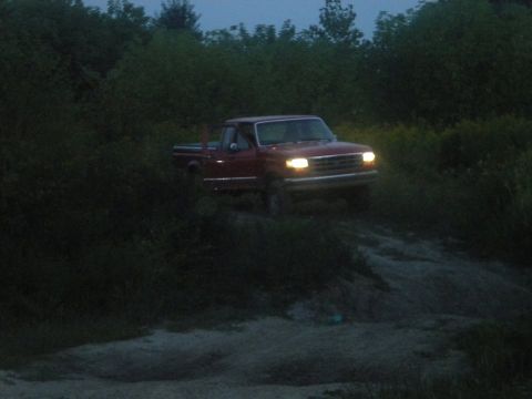 1995 Ford F-150 - A red pickup truck navigates a dirt path surrounded by foliage as twilight deepens.
