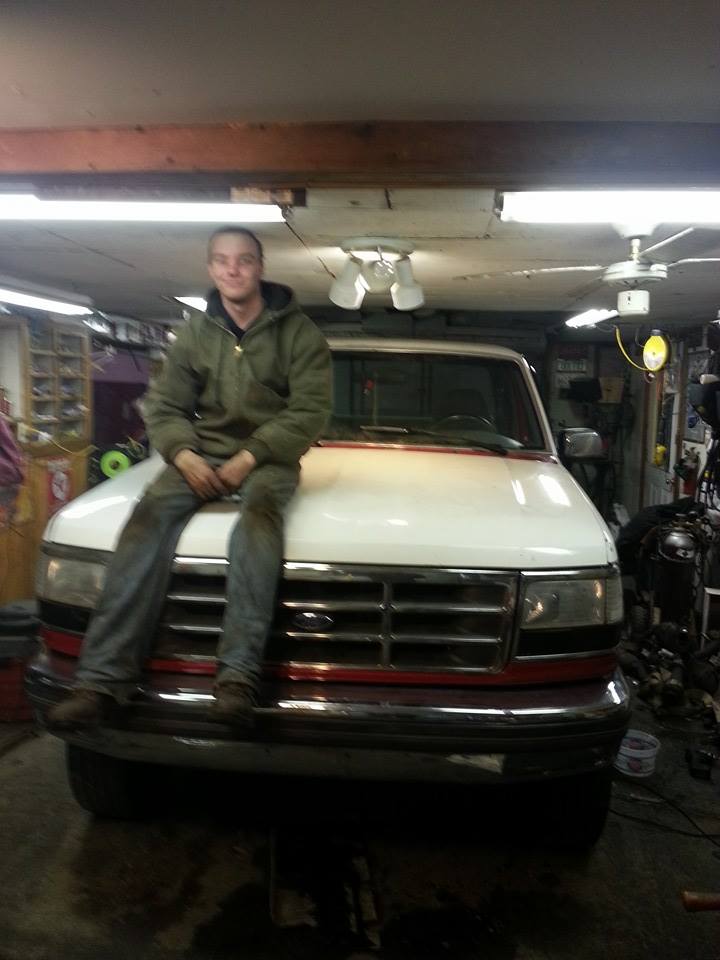 Man in green jacket and jeans sits on the hood of a white 1995 Ford F-150 truck in a garage