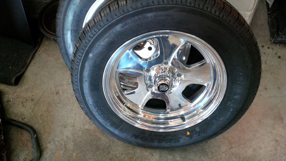 Two polished chrome slotted wheels with all-season radial tires leaning against a garage wall