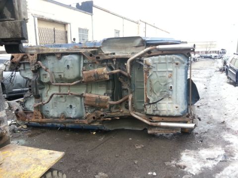 2004 Mercury Grand Marquis - The underside of a heavily rusted blue car chassis is visible, flipped upside down in a junkyard setting.