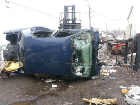 2004 Mercury Grand Marquis - A severely damaged blue car body is being lifted by a forklift in a junkyard.
