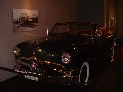 1999 Ford Crown Victoria - A classic dark green convertible is displayed indoors, with a black and white photo on the wall behind it.