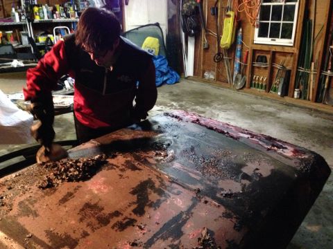 1979 Ford Bronco - A person is actively removing rust and debris from a car hood in a cluttered garage setting.