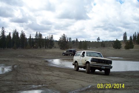 1990 Ford Bronco - Two SUVs drive through a muddy, puddle-filled area with trees in the background under a cloudy sky.