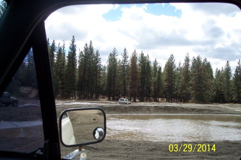 1990 Ford Bronco - View from inside a vehicle shows a white SUV on a dirt track with trees and water.