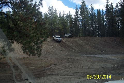 1990 Ford Bronco - Two trucks navigate a steep dirt incline surrounded by pine trees under a cloudy sky.