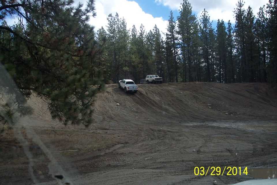 Two trucks driving up a dirt hill in a wooded area. One is white, the other is white