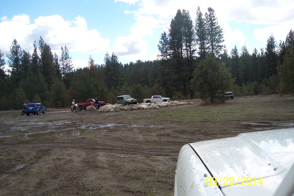 Several off-road vehicles, including a blue SUV and red truck, are in a muddy area with trees in the background