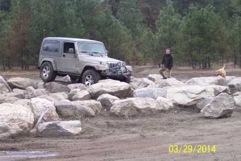1990 Ford Bronco - A silver Jeep Wrangler navigates a rocky terrain, with a man and a dog observing the scene.