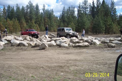 1990 Ford Bronco - Two off-road vehicles navigate a rocky obstacle course surrounded by pine trees.