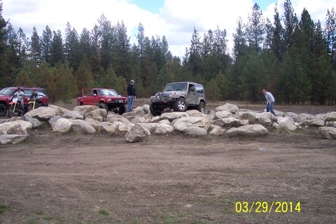 1990 Ford Bronco - Several off-road vehicles navigate a rocky obstacle course with a wooded background.