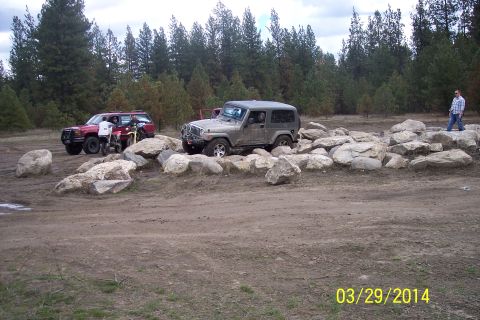 1990 Ford Bronco - A gray Jeep Wrangler maneuvers through a challenging rock obstacle course, with a red SUV and dirt bike visible in the background.