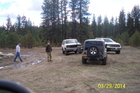 1990 Ford Bronco - Several vehicles are parked in a dirt lot surrounded by trees, with two men standing nearby.