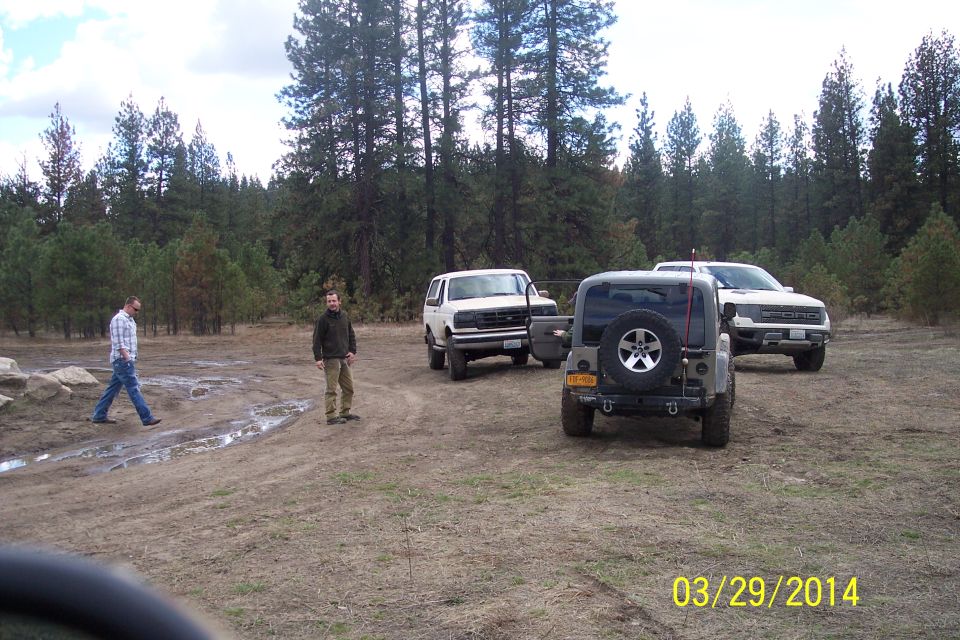 Three SUVs and two guys in a dirt lot with trees behind them. One SUV is a white Ford Bronco, another is a gray Jeep, and the third is a white pickup truck
