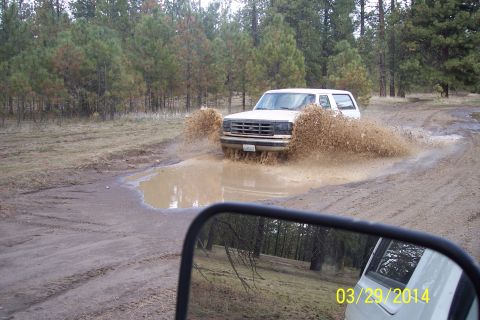 1990 Ford Bronco - A white SUV kicks up a large spray of mud as it drives through a puddle on a dirt road.