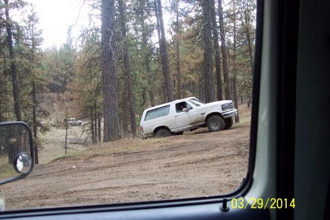 1990 Ford Bronco - A white Ford Bronco navigates a muddy off-road trail through a forest, with another vehicle visible in the distance.