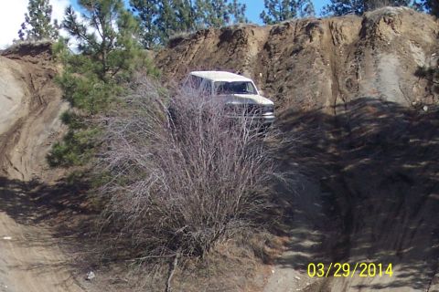 1990 Ford Bronco - A white SUV navigates a steep, muddy incline on a dirt trail, surrounded by sparse vegetation and trees.