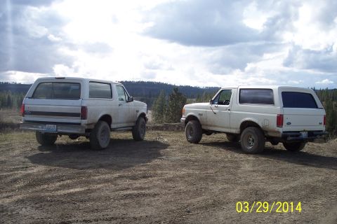 1990 Ford Bronco - Two white Ford Broncos are parked on a dirt road with a wooded hillside in the background. The vehicle on the right is likely a 1992 model.