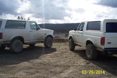 1990 Ford Bronco - Two white Ford Broncos are parked on a dirt lot with a wooded hillside under a cloudy sky.