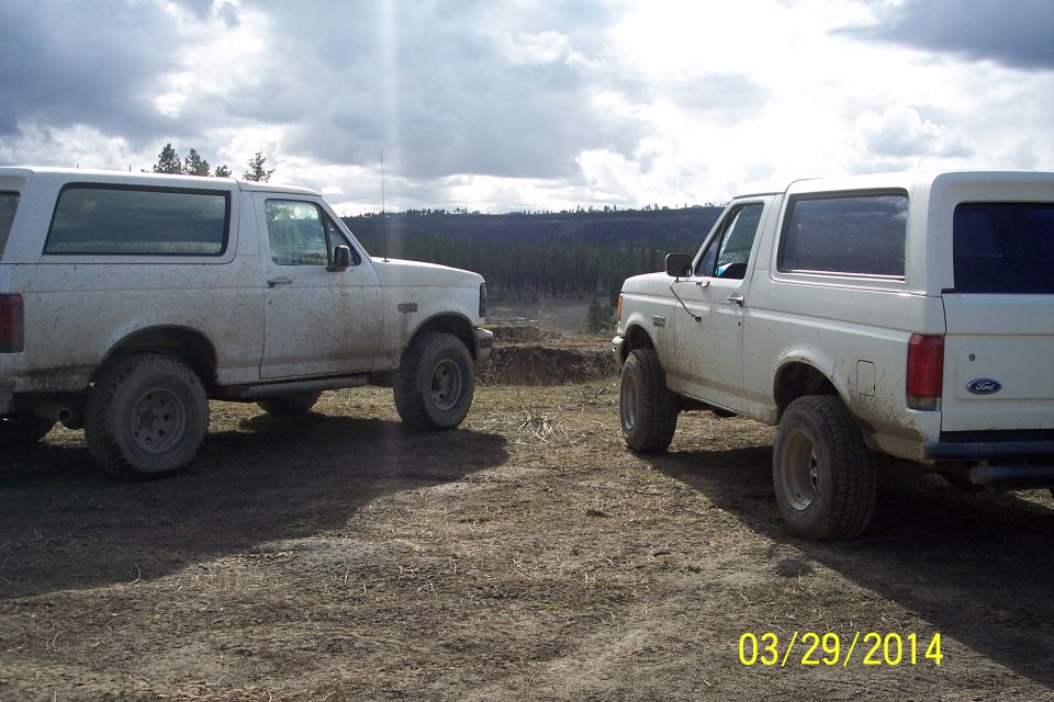 Two white Ford Broncos parked on dirt with trees and cloudy sky in background. One Bronco is in the foreground, facing right. The other is behind it, facing left