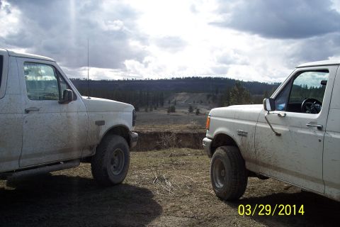 1990 Ford Bronco - Two white Ford Broncos are parked on a dirt road, facing each other under a cloudy sky.