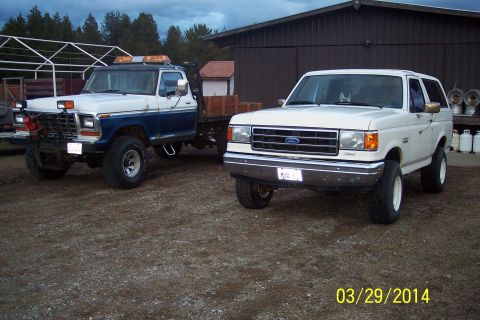 1990 Ford Bronco - Two trucks, a white Ford Bronco and a blue/white tow truck, are parked outside a building.