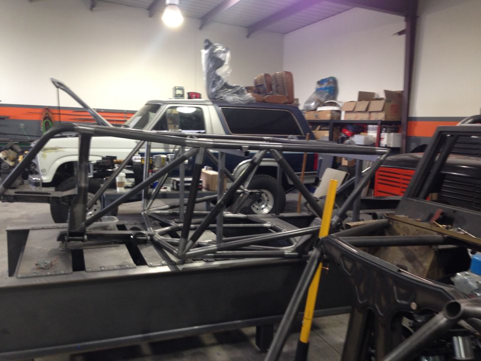 Ford Bronco in a workshop, partially obscured by a custom roll cage structure. Seats are visible in the background