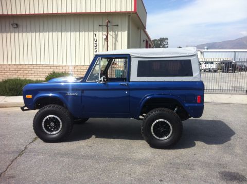 1986 Ford Bronco - A blue Ford Bronco with a grey soft top is parked on asphalt in front of a building.