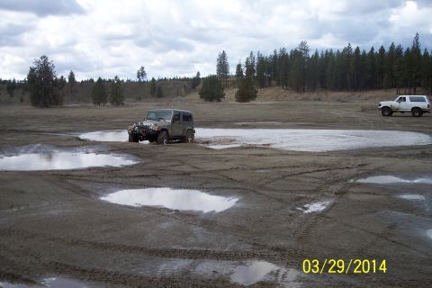 2005 Jeep Wrangler - Two off-road vehicles navigate a muddy terrain with scattered puddles and trees in the background.
