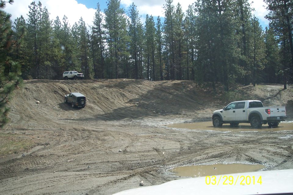 Three off-road vehicles, including a white pickup truck and two SUVs, on a muddy dirt track with trees in the background