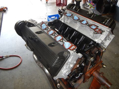 2003 Ford Crown Victoria - Close-up of a V8 engine showing a finned black valve cover, exposed cylinder bank, and blue-taped intake ports during assembly on a stand.