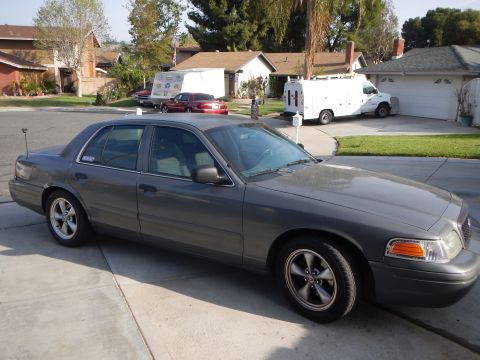 2003 Ford Crown Victoria - Front three-quarter view of a gray full-size Ford sedan parked on a driveway in a suburban neighborhood.