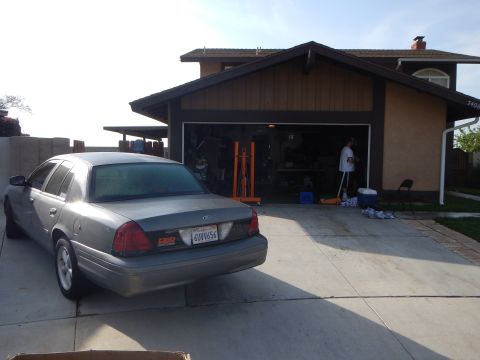 2003 Ford Crown Victoria - Rear three-quarter view of a gray Ford sedan with a California plate parked outside an open suburban garage.