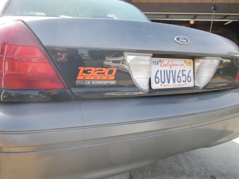 2003 Ford Crown Victoria - Close-up of a gray Ford sedan's rear trunk lid with a Ford emblem, California plate 6UVV656, and a 1320 Video Police Interceptor sticker.