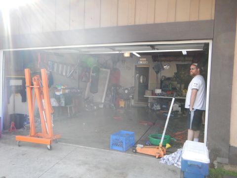 2003 Ford Crown Victoria - bearded man stands at the entrance of a cluttered garage holding a hose, with an orange engine hoist and tools inside.