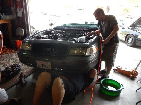 2003 Ford Crown Victoria - Two men work on the engine of a gray Ford sedan with its hood up in a garage while another person lies underneath the front bumper.