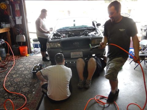 2003 Ford Crown Victoria - Three men gather around the front of a gray Ford sedan with its hood open in a garage, one holding an orange extension cord.