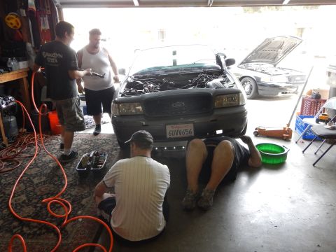 2003 Ford Crown Victoria - Several men work around the open hood of a gray Ford sedan in a garage, with a green drain pan and floor jack on the floor.
