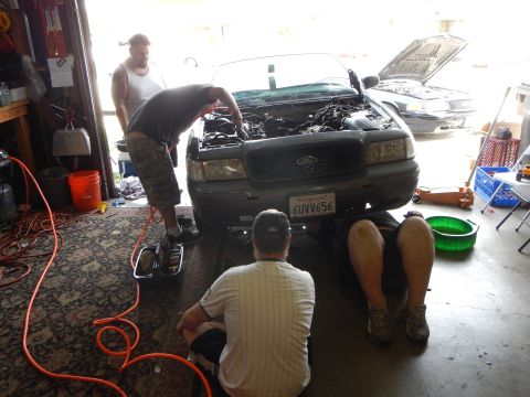 2003 Ford Crown Victoria - Men work on the engine of a gray Ford sedan with its hood raised in a garage, surrounded by tools and orange extension cords.