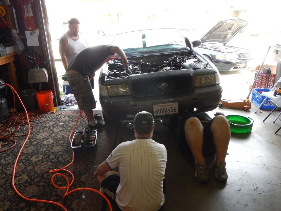 Men working under the hood of a gray Ford sedan in a garage, plate 6UVV656