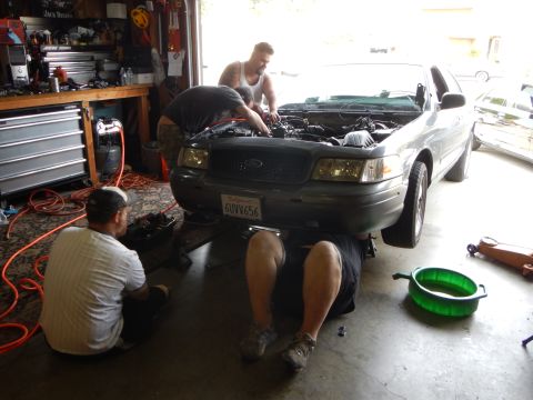 2003 Ford Crown Victoria - Two men work on a gray Ford sedan with the hood up in a garage as a green drain pan sits on the floor beside them.