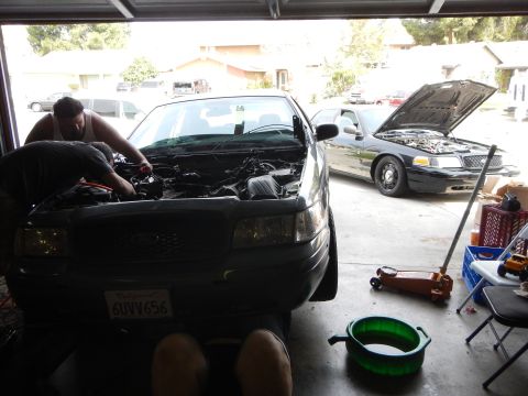 2003 Ford Crown Victoria - gray Ford sedan with its hood up sits in a garage being worked on, with a second gray Ford parked in the driveway behind it.