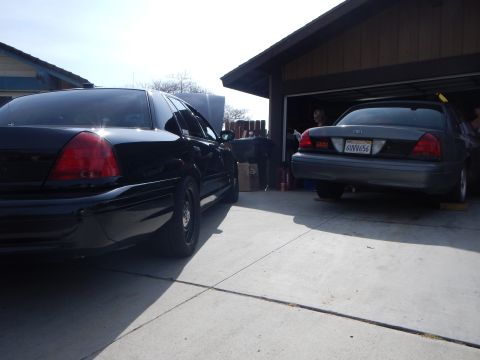 2003 Ford Crown Victoria - black Ford sedan sits in a driveway next to a garage holding a gray Ford sedan with California plate 6UVV656.