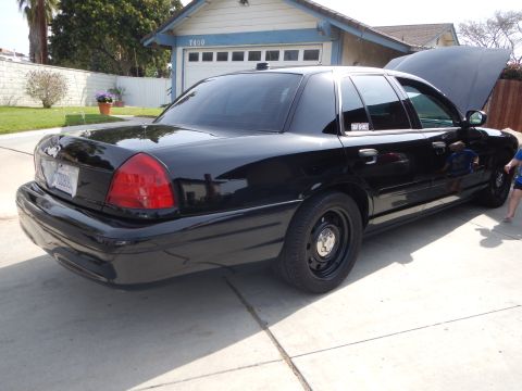 2003 Ford Crown Victoria - Rear three-quarter view of a black Ford Crown Victoria with black steel wheels and its hood up, parked in a driveway by a blue house.